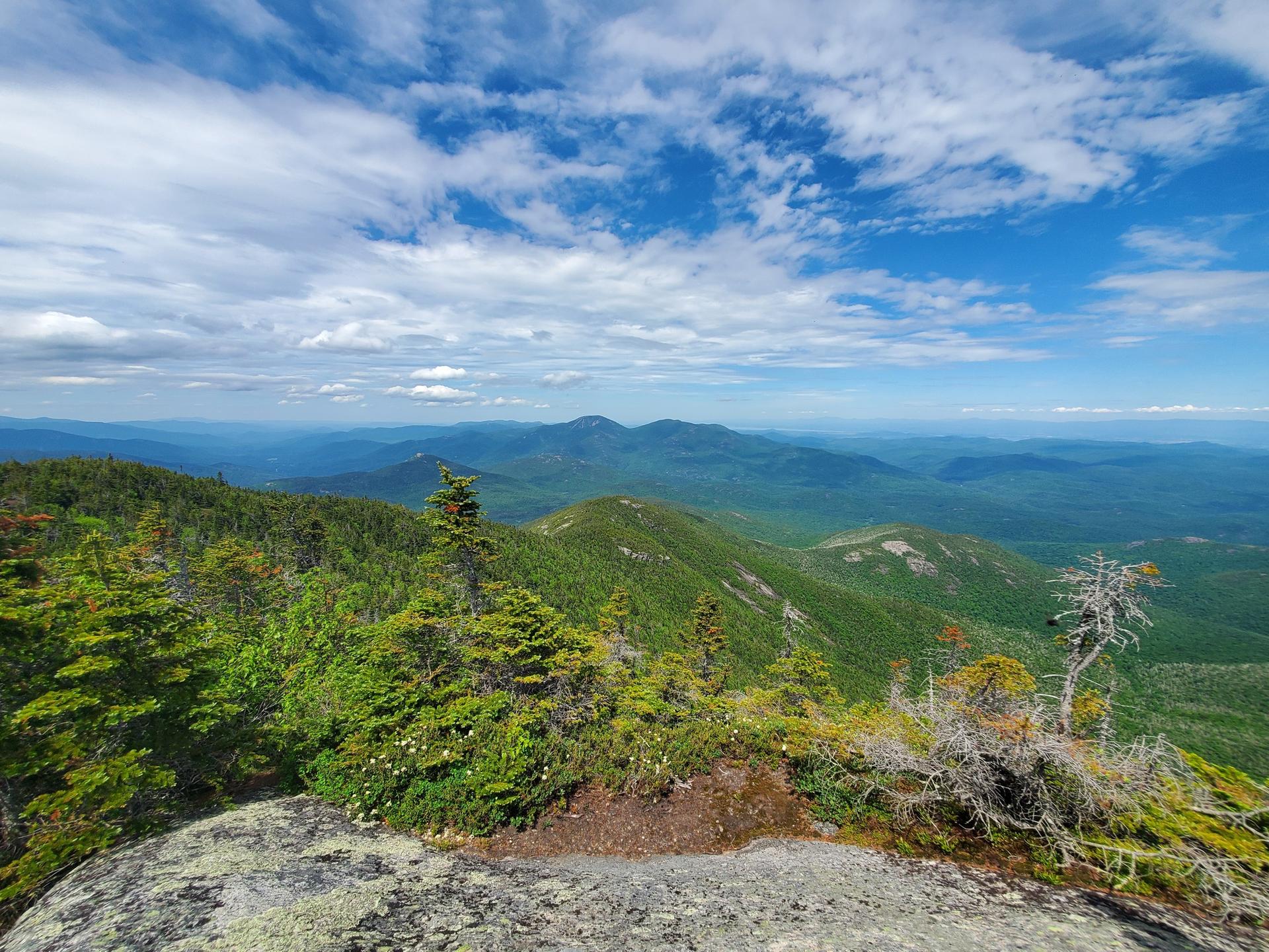 Vast green mountain ranges under a partly cloudy blue sky from a rocky viewpoint.