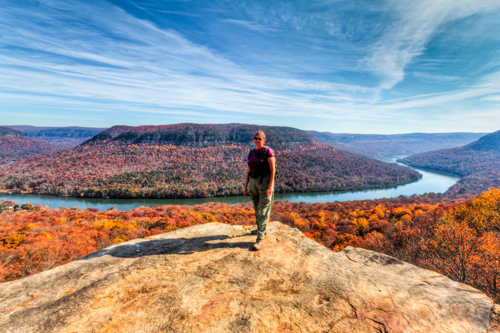 Person stands on a rocky overlook, viewing a river winding through autumn hills.