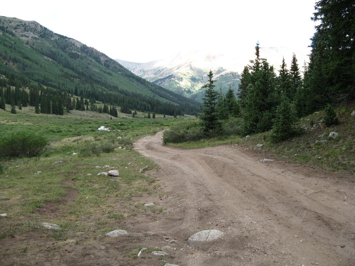 A winding dirt road through a green valley with pine trees and distant mountains.