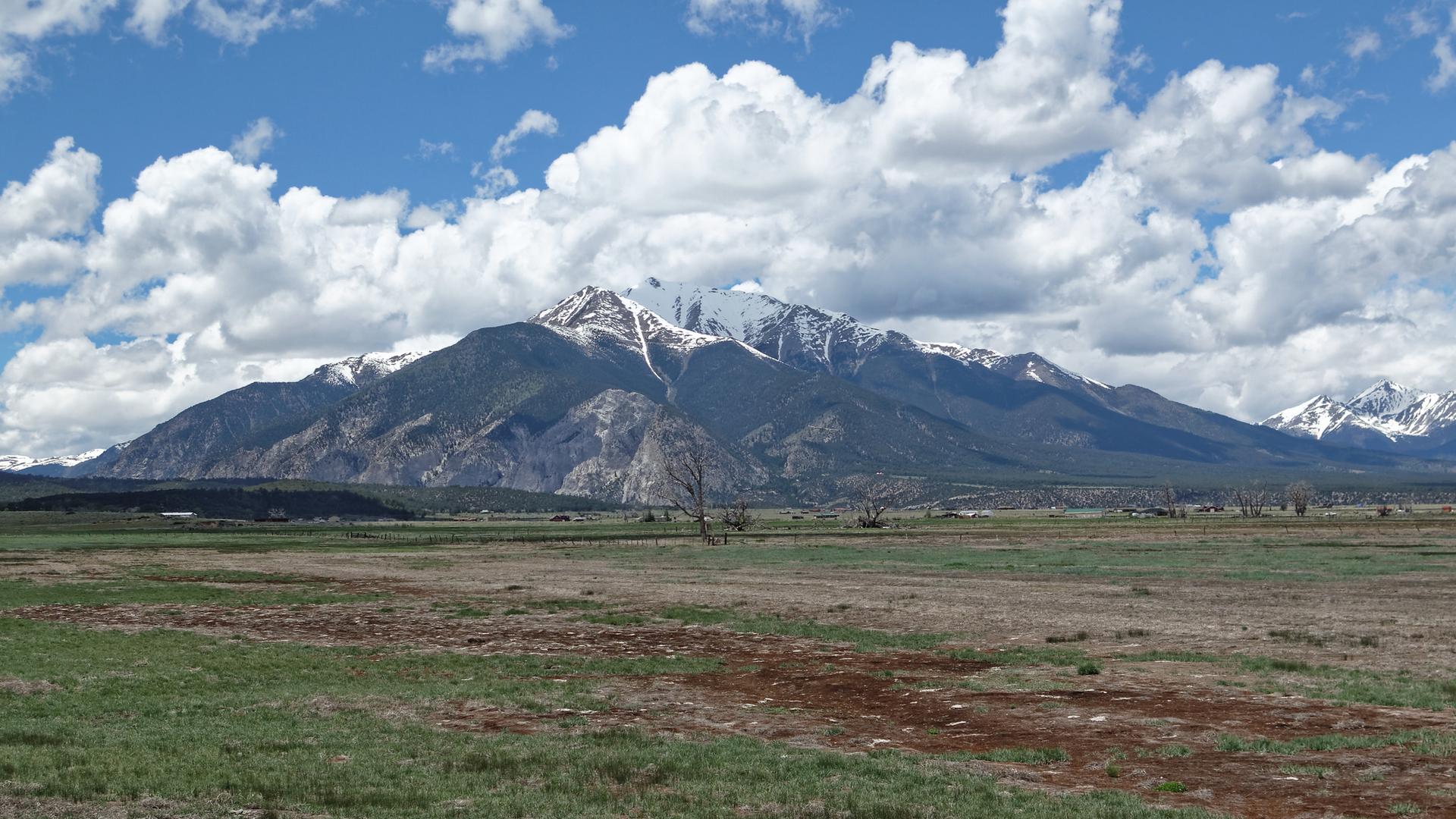 Expansive field with distant snow-capped mountains under a dramatic cloudy sky.