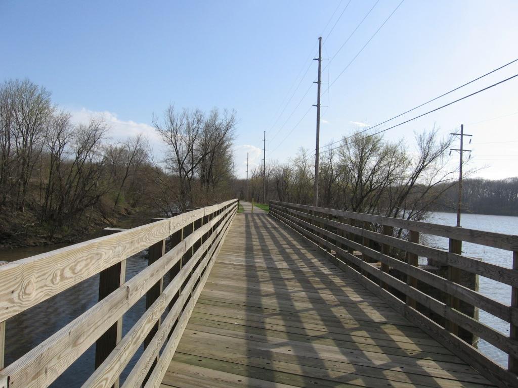 Long wooden boardwalk over water, bare trees, and utility poles under bright sun.