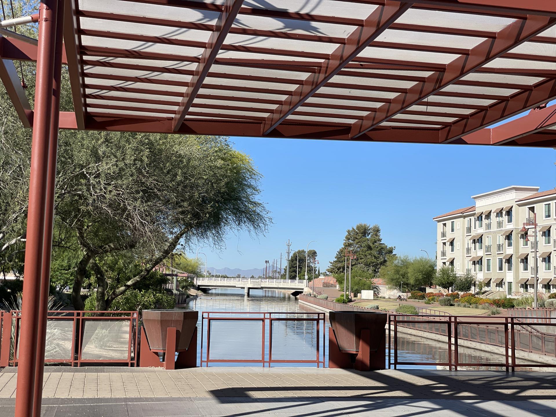View through a pergola of a canal, bridge, and distant building on a sunny day.