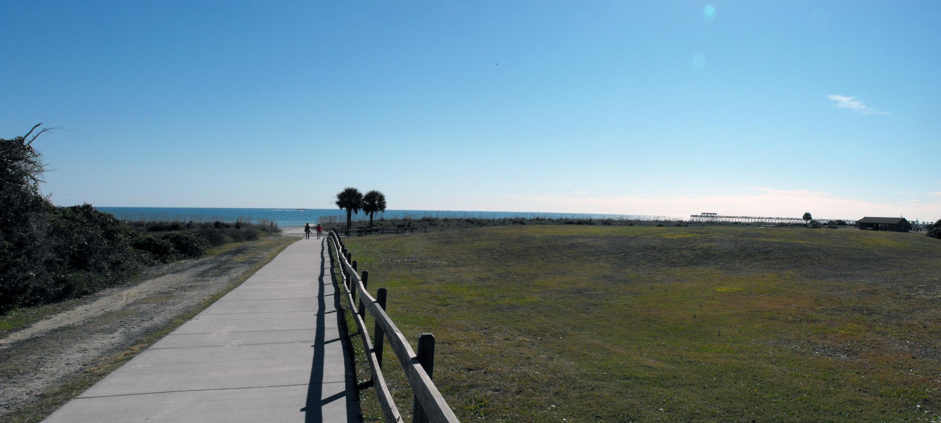 Paved path leading to a sunny beach with palm trees and ocean.