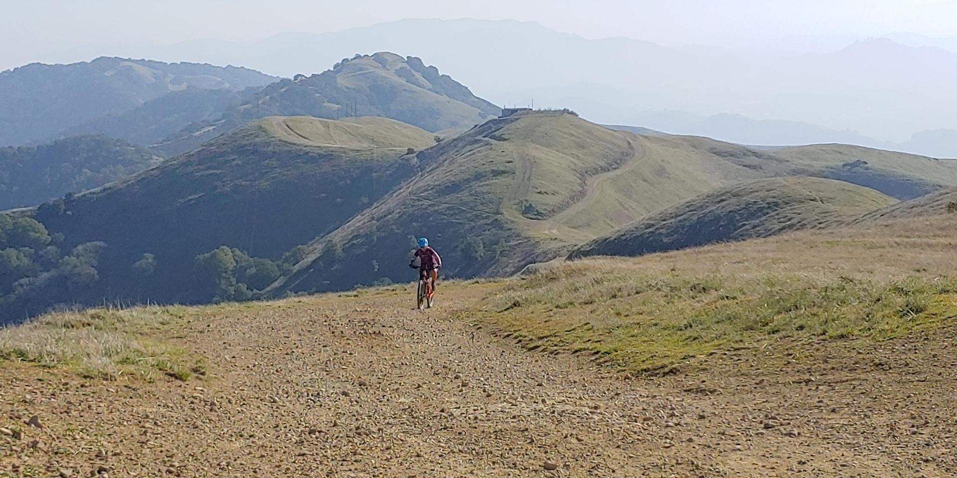 A person on a bicycle rides a dirt path through rolling grassy hills.
