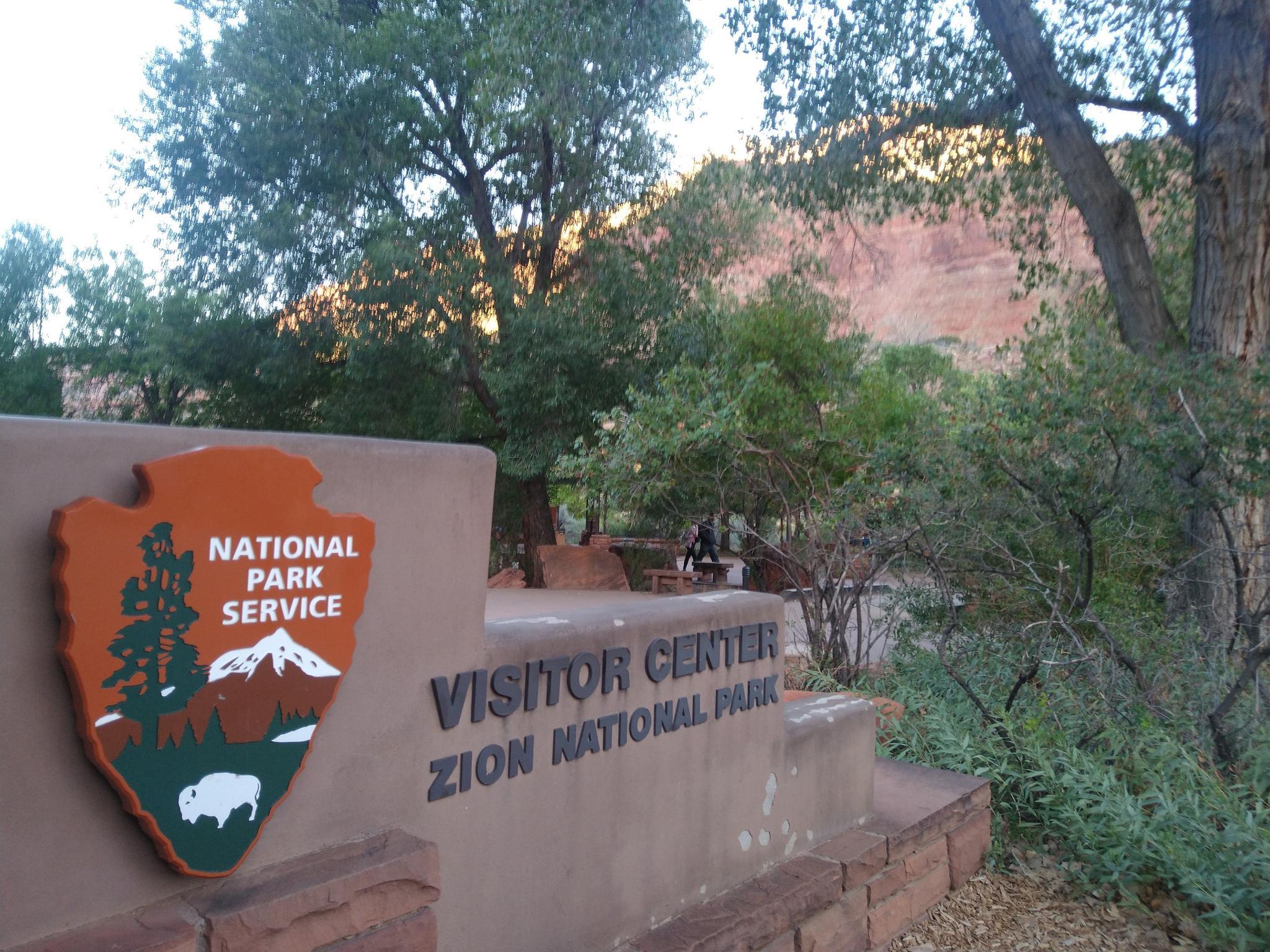 A park service sign for a visitor center, surrounded by trees and distant cliffs.