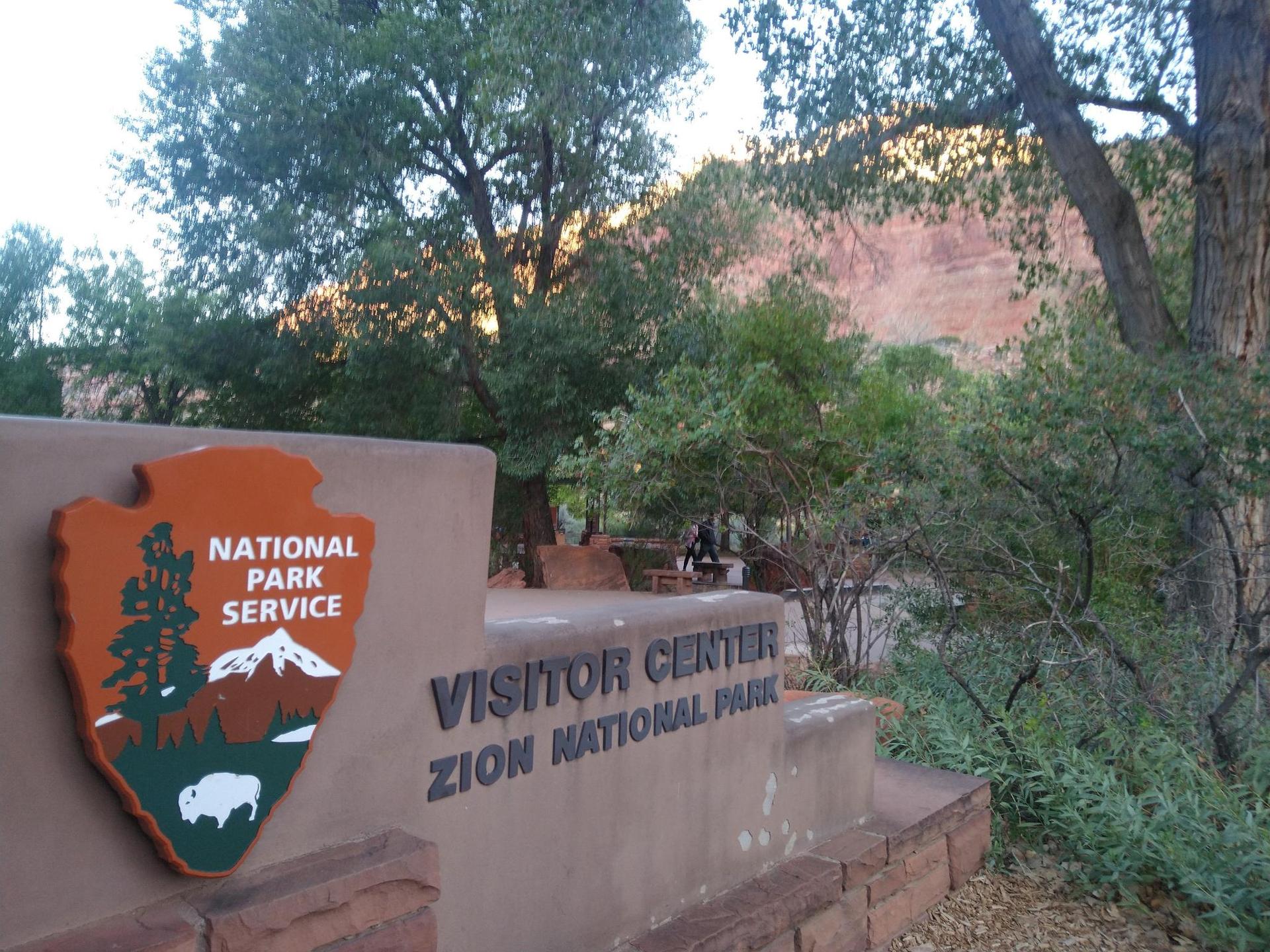 A park service sign for a visitor center, surrounded by trees and distant cliffs.