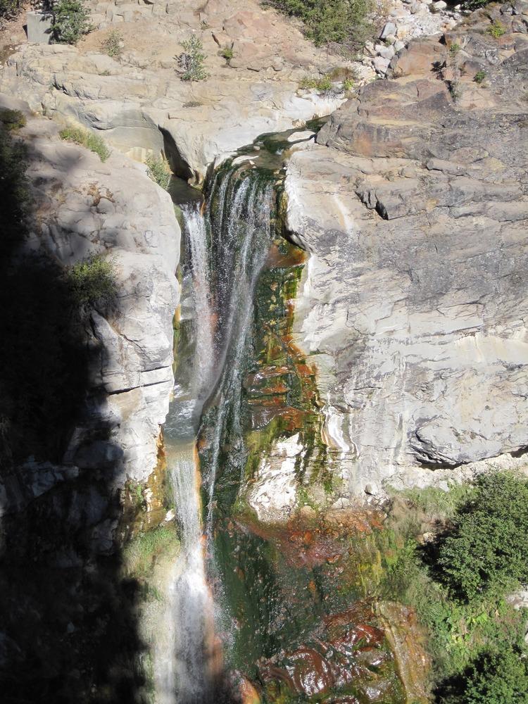 A narrow waterfall cascades down a colorful, mossy rock face in sunlight.