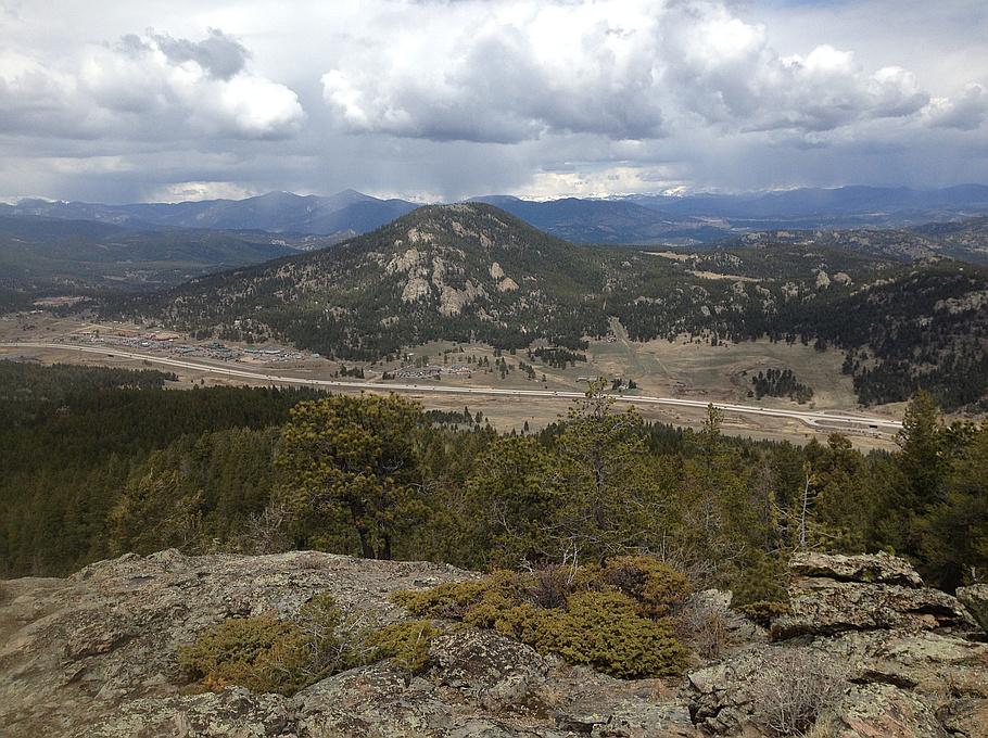 Rocky foreground overlooks a forested valley, road, and distant mountains under cloudy sky.