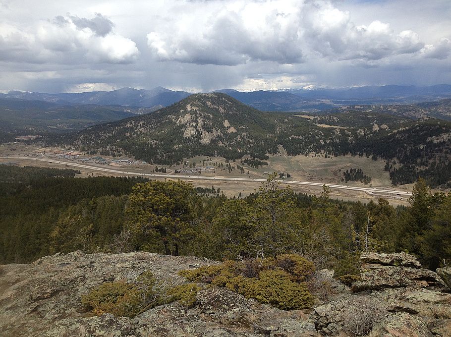 Rocky foreground overlooks a forested valley, road, and distant mountains under cloudy sky. - Legault Mountain loop from Aspen Park