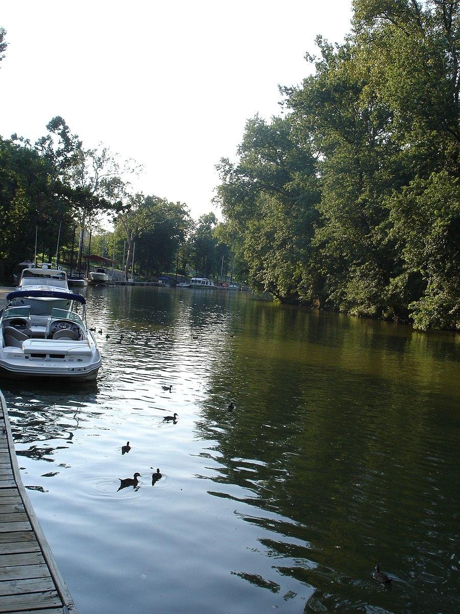 Boats docked on a narrow, tree-lined waterway with ducks swimming in bright light.