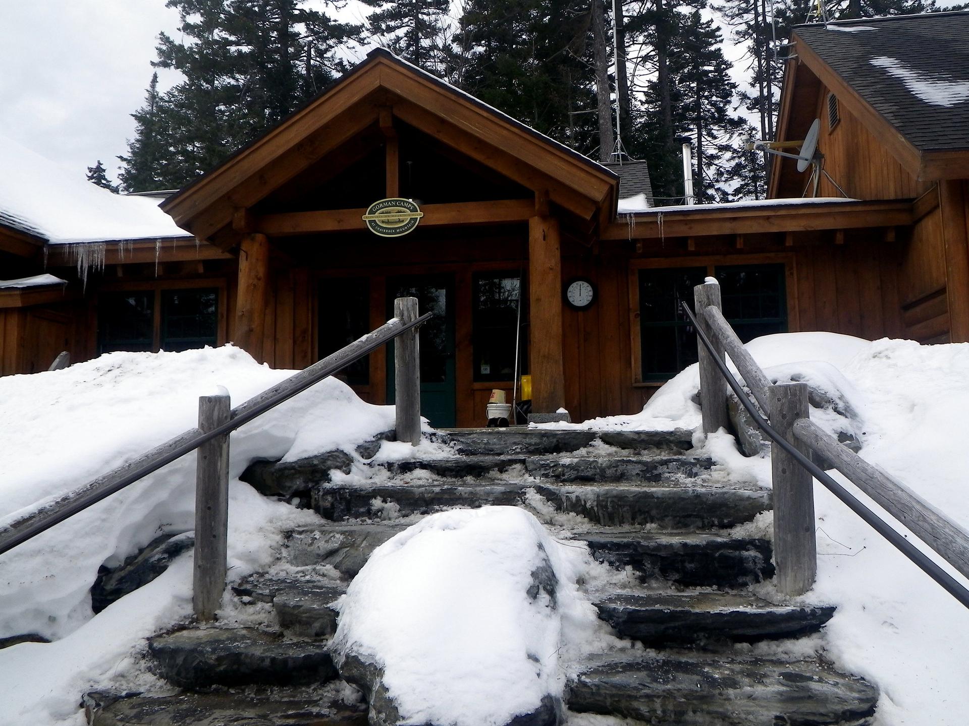 Snow-covered stone steps lead to a rustic wooden lodge entrance on an overcast day.