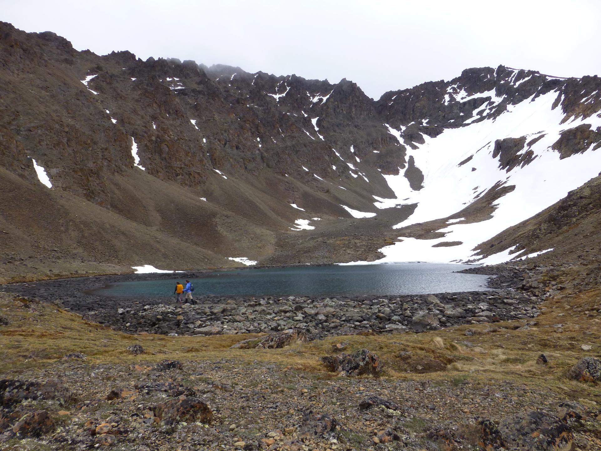 Two hikers by a rocky mountain lake under an overcast sky.