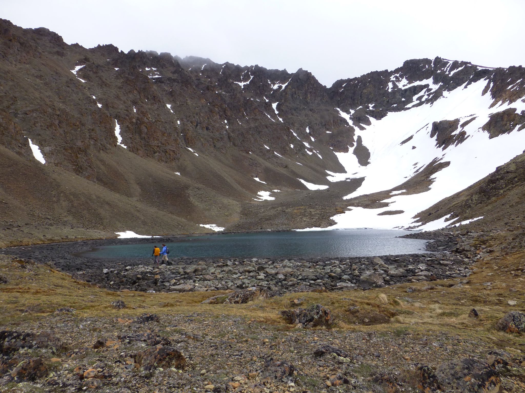 Two hikers by a rocky mountain lake under an overcast sky. - Hidden Lake Trail