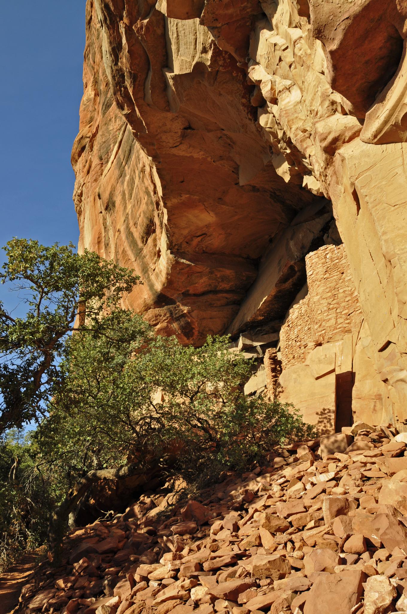 Ancient stone dwelling built into a red rock cliff with green trees.