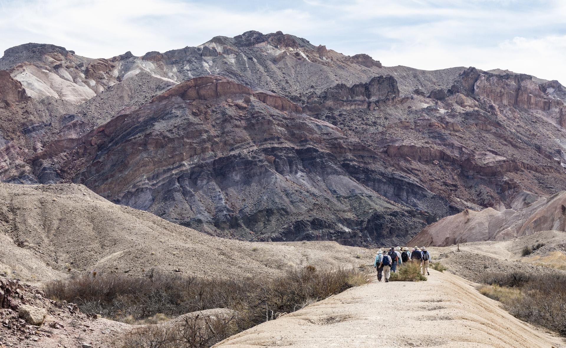 Hikers on a dirt path with vast, colorful, rugged mountains.
