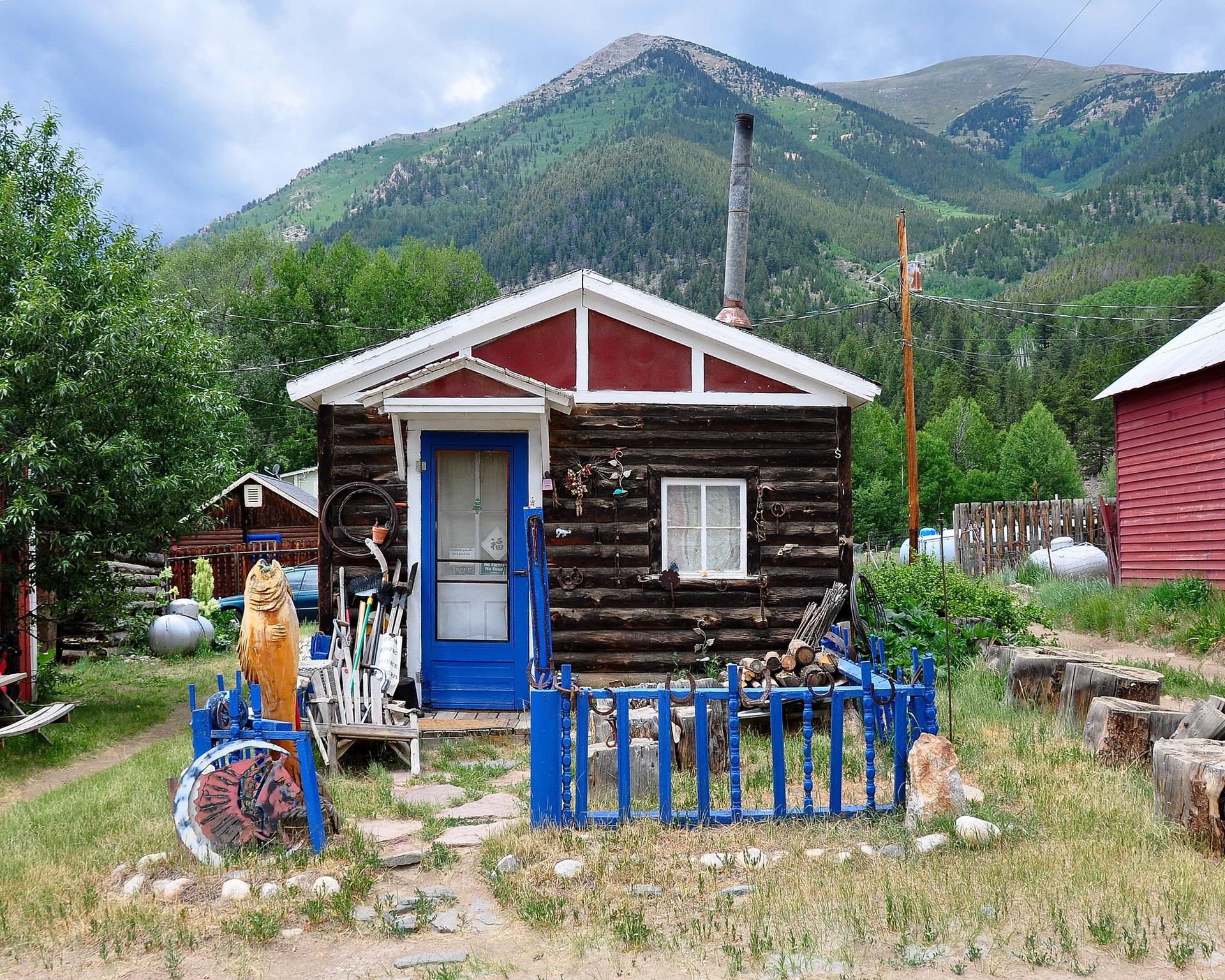A rustic log cabin with a red roof and blue door, mountains behind.