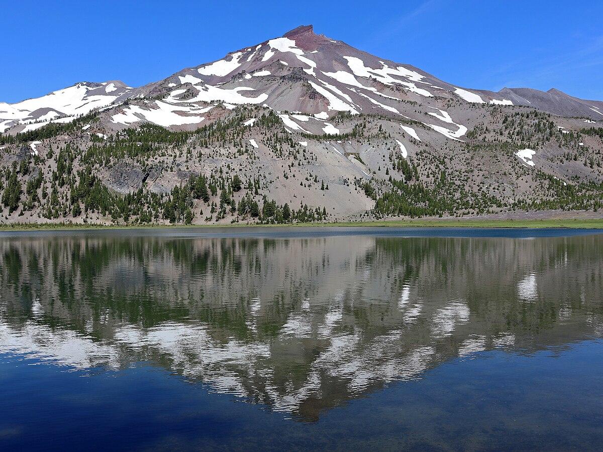 Snow-capped mountain reflecting in a calm lake under a bright blue sky.