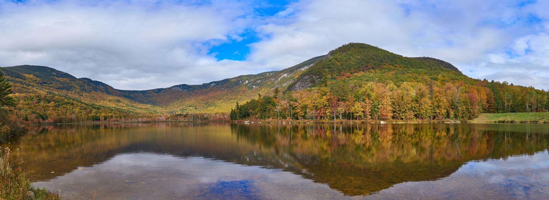 Calm lake reflecting autumn-colored mountains and trees under a cloudy sky.