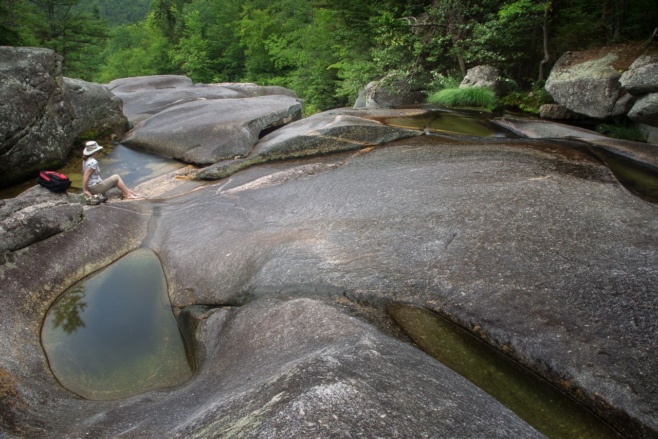 Person sitting on sunlit, smooth rocks next to clear water pools in a forest. - Step Falls Trail