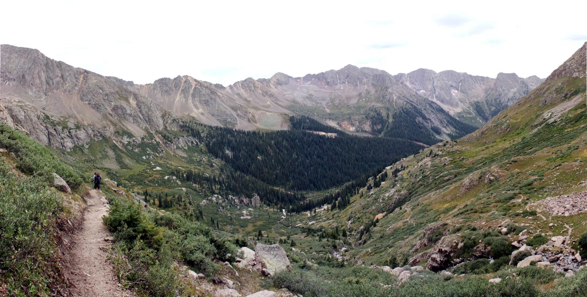Hiker on a dirt trail overlooking a vast mountain valley with forests under cloudy skies.