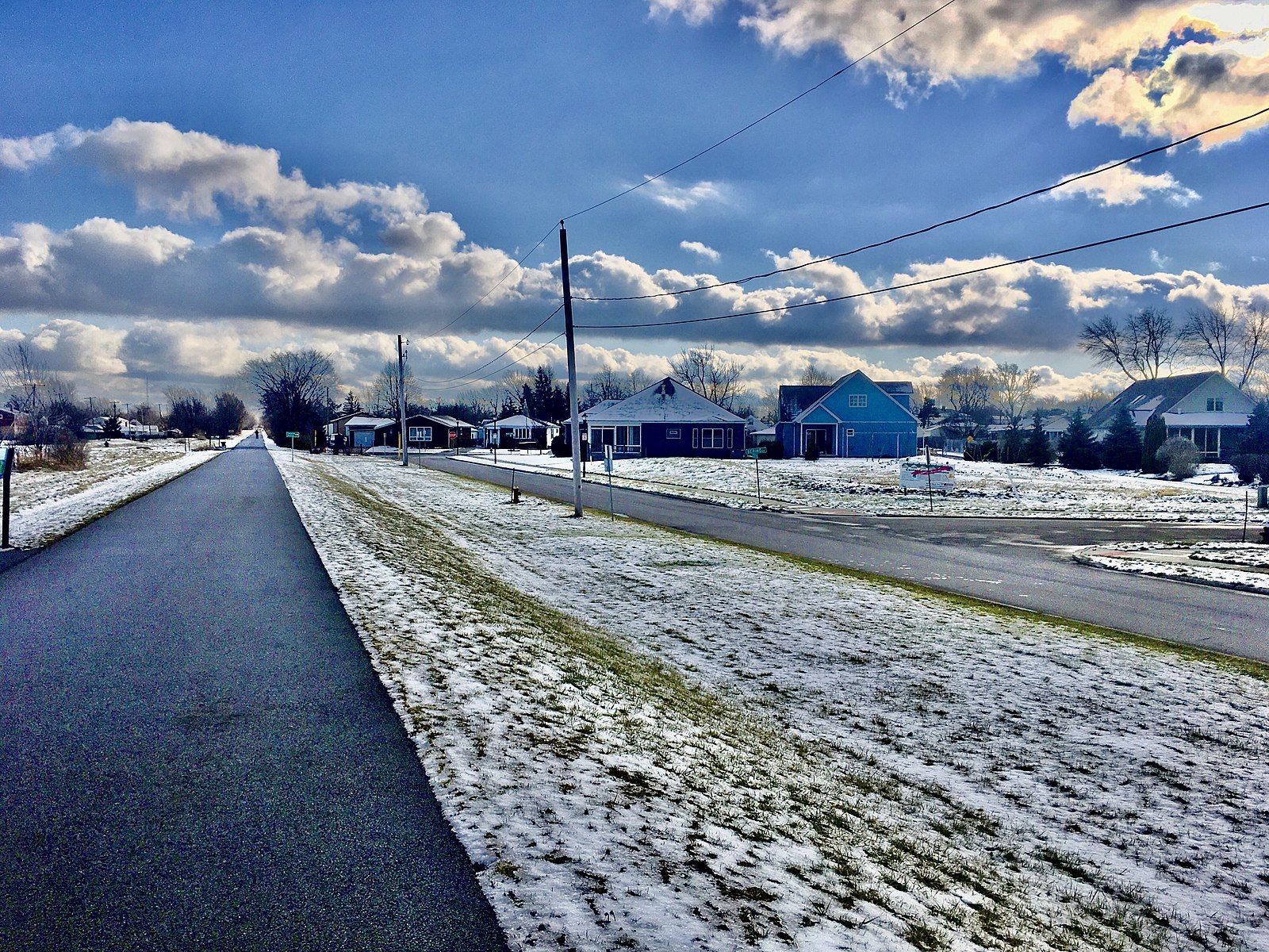 A snow-dusted road and grassy area with houses under a bright, cloudy sky. - Clarence Akron Peanut Line Bike Pathways and Tonawanda Rails-to-Trails loop