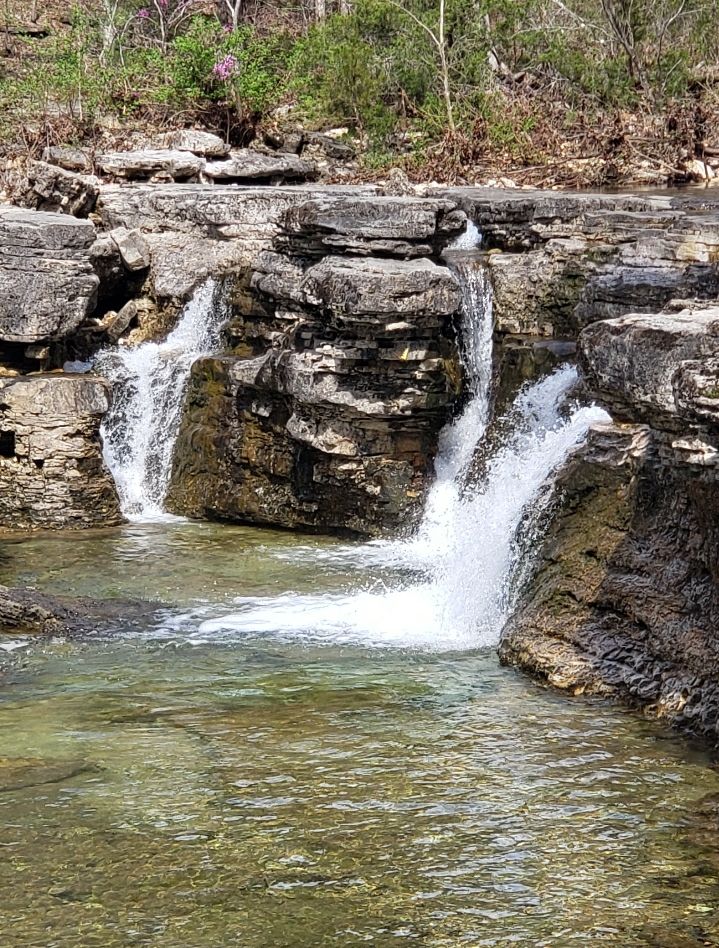 Waterfalls cascade over layered rocks into a clear stream under bright daylight. - Hercules Glade Backpacking Loop