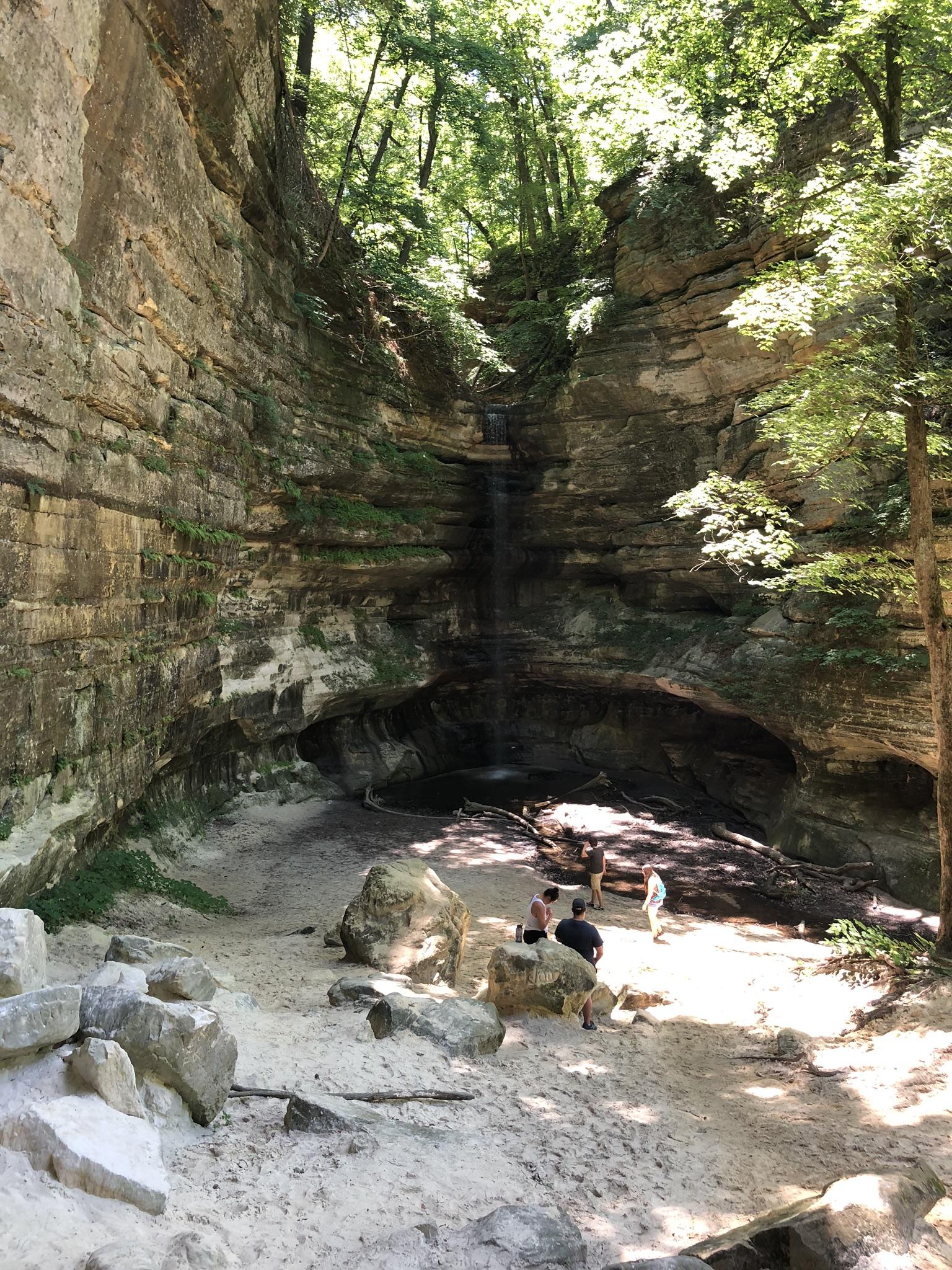 People gathered at the base of a waterfall in a sunny, green canyon.