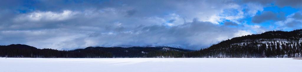 Panoramic view of a snowy field with dark forested mountains under a cloudy sky.