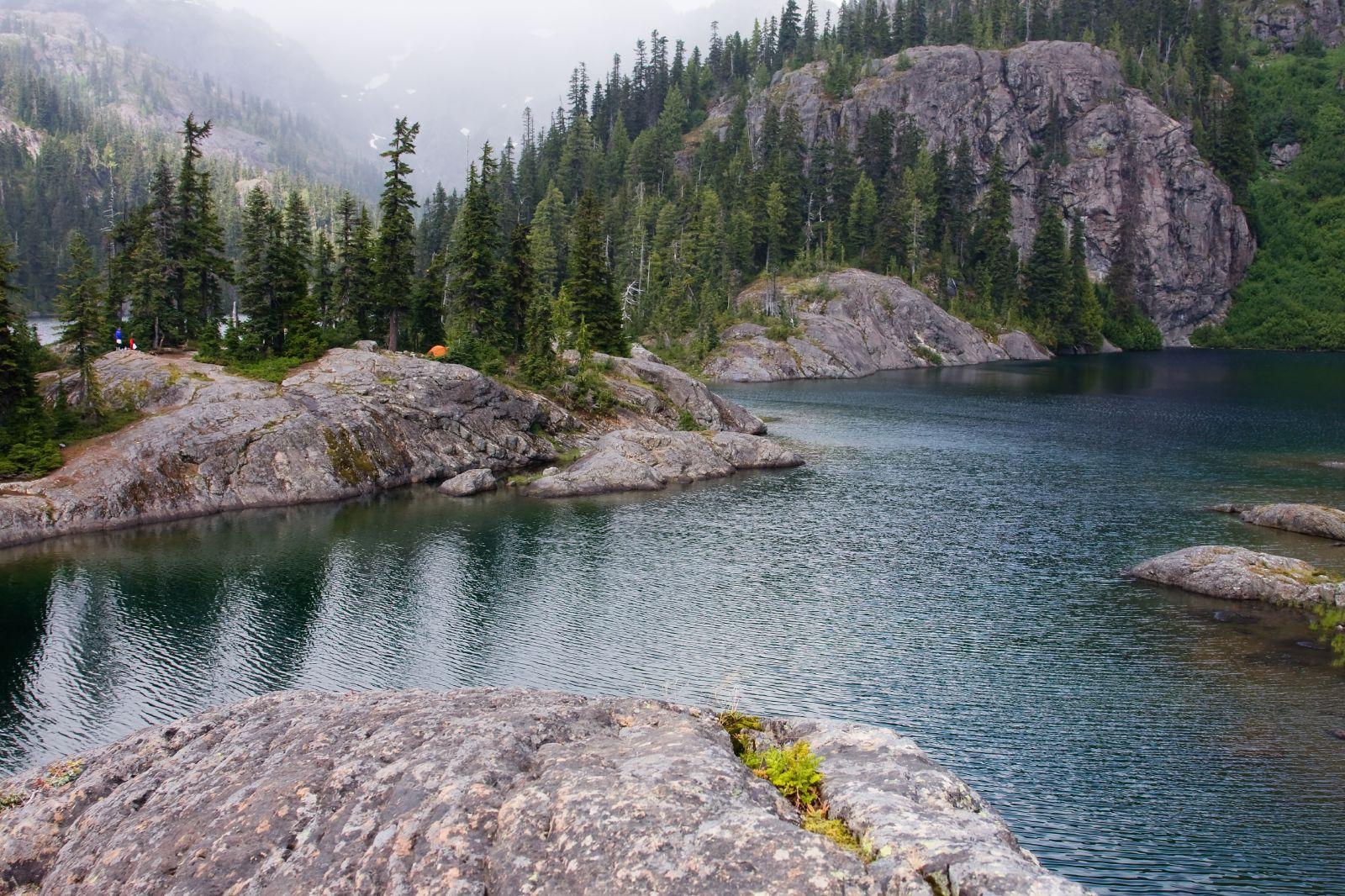 Rocky lake shore with dark water, pine trees, and misty mountains.