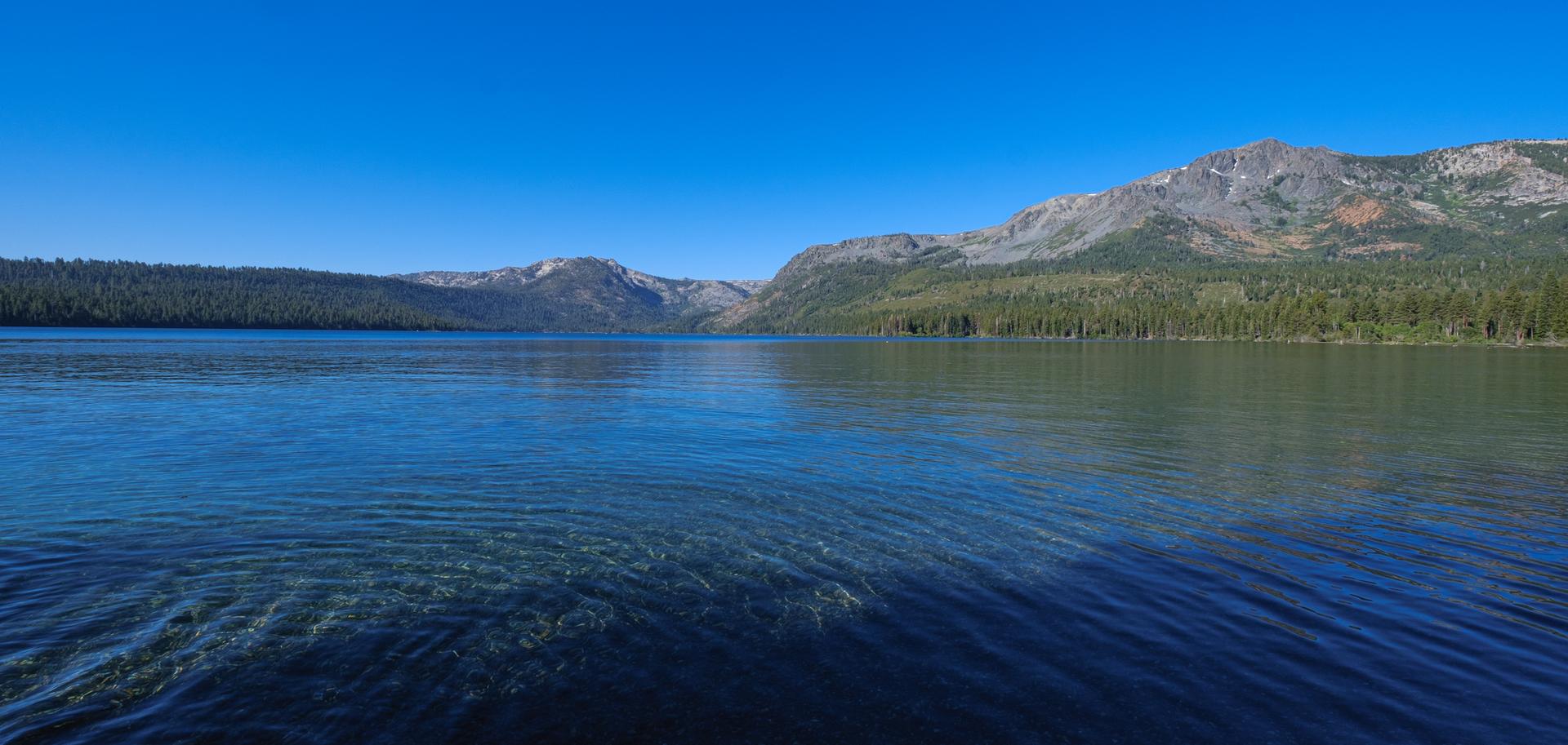 Clear blue lake with forested shores and majestic mountains under a bright sky.