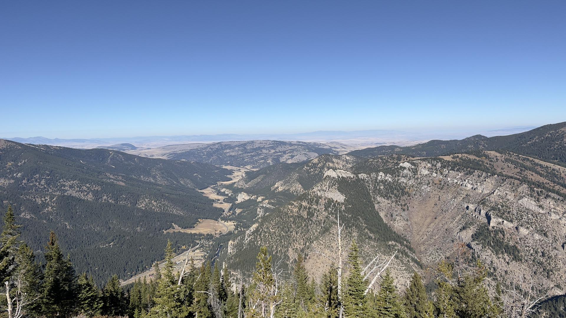 Expansive mountain vista with forested slopes, a valley, and clear blue sky.