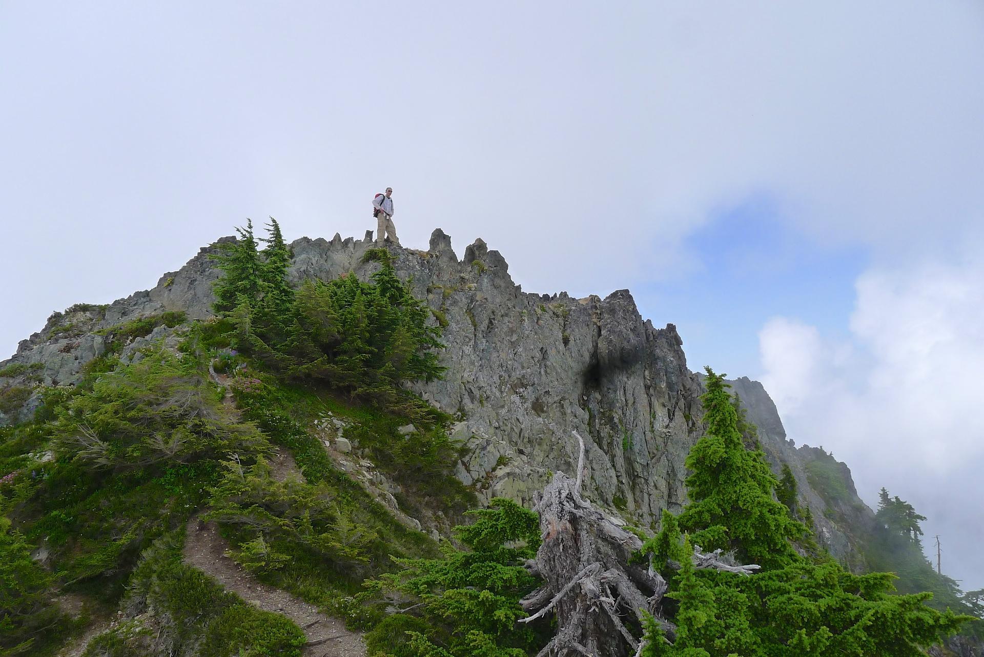 Person stands atop a rugged, rocky mountain peak under cloudy skies.