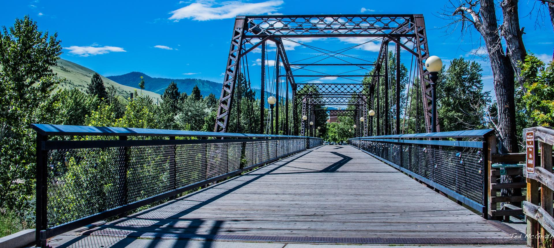 A long metal truss bridge with a wooden deck under a bright blue sky.