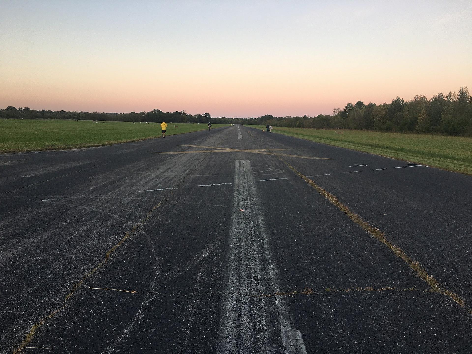 Long asphalt runway with faded markings, surrounded by green fields at dusk.