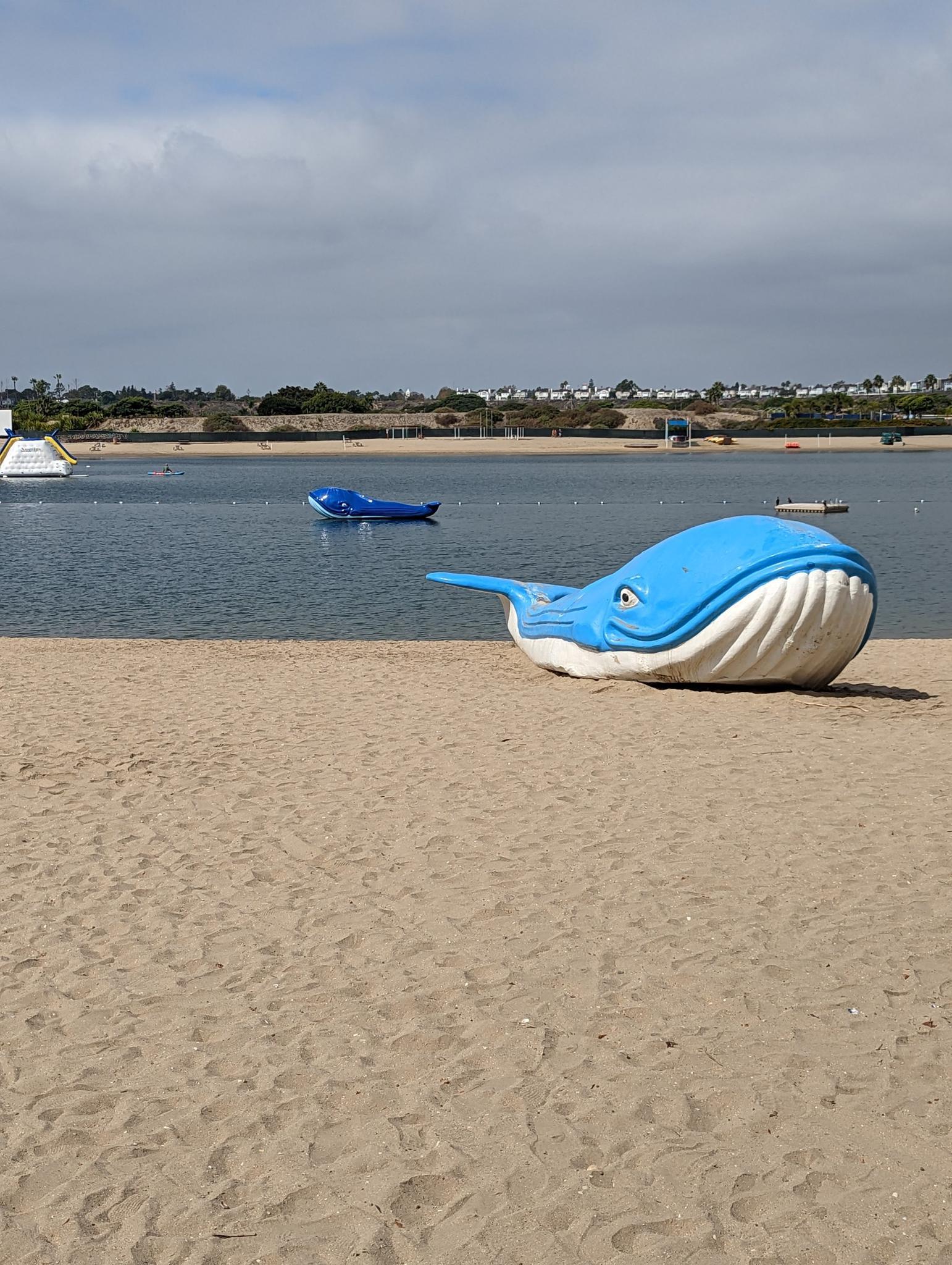 Two blue whale sculptures on a sandy beach by calm water.
