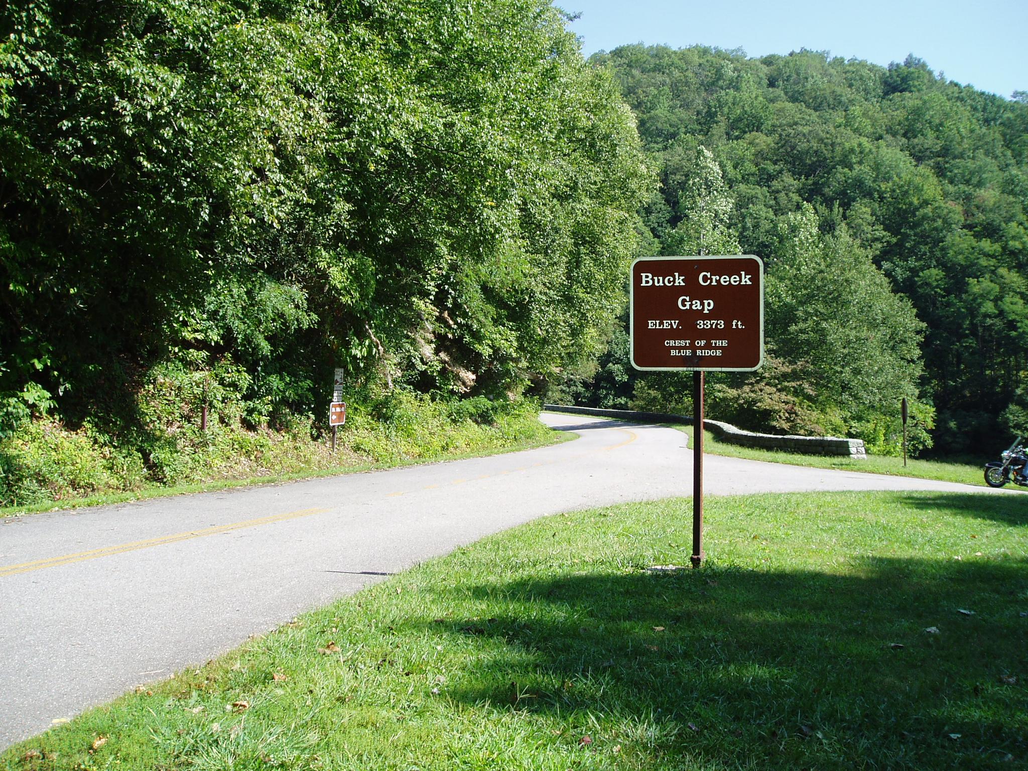 Sunny roadside with a brown sign, paved road, green forested hills, and a distant motorcycle.