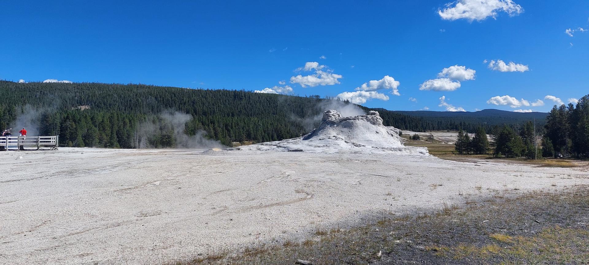 A panoramic view of a steaming geyser cone in a vast thermal field under a blue sky.