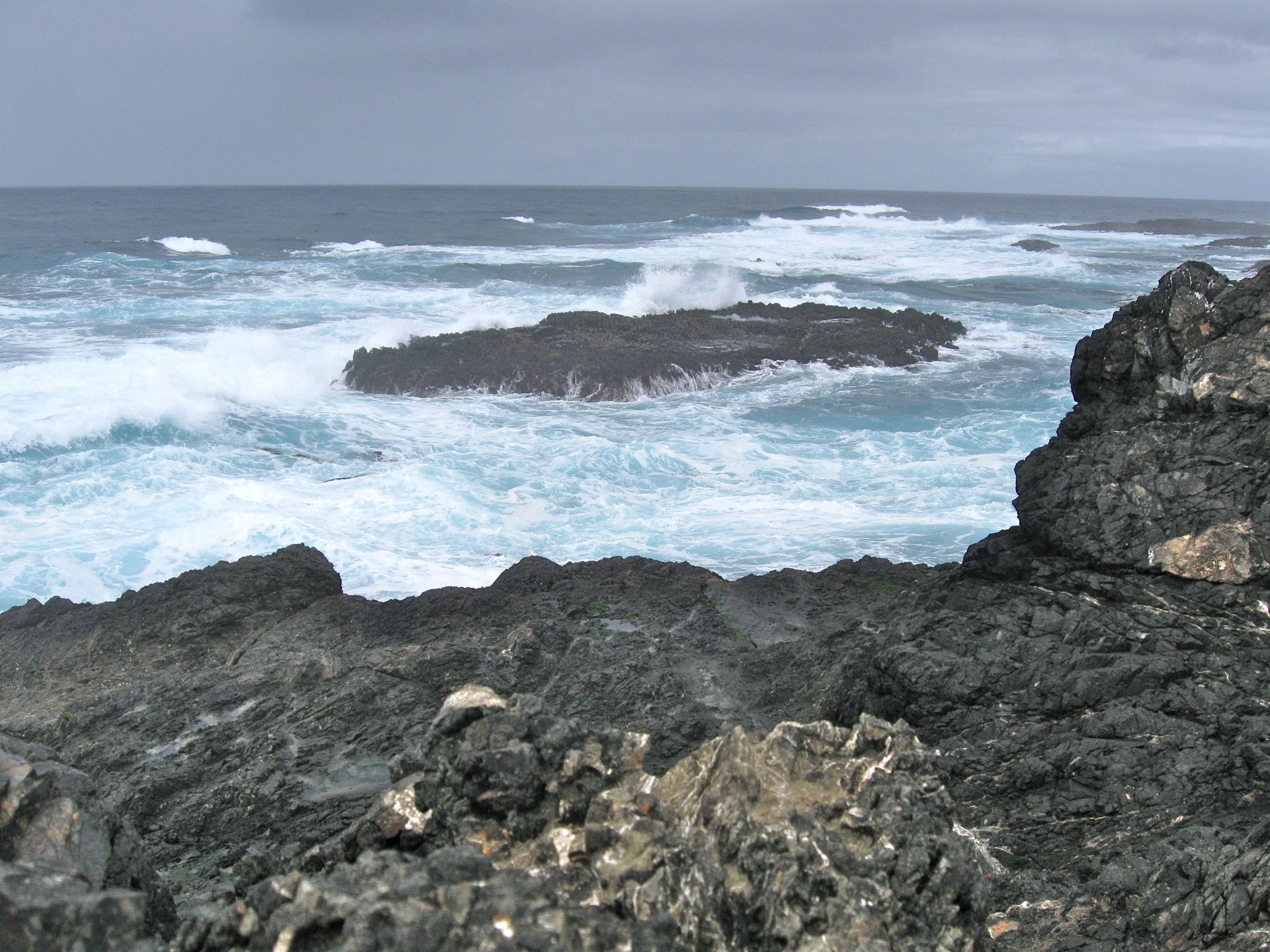 Turbulent ocean waves crash against dark, rugged volcanic rocks under an overcast sky. - Spring Ranch Trail