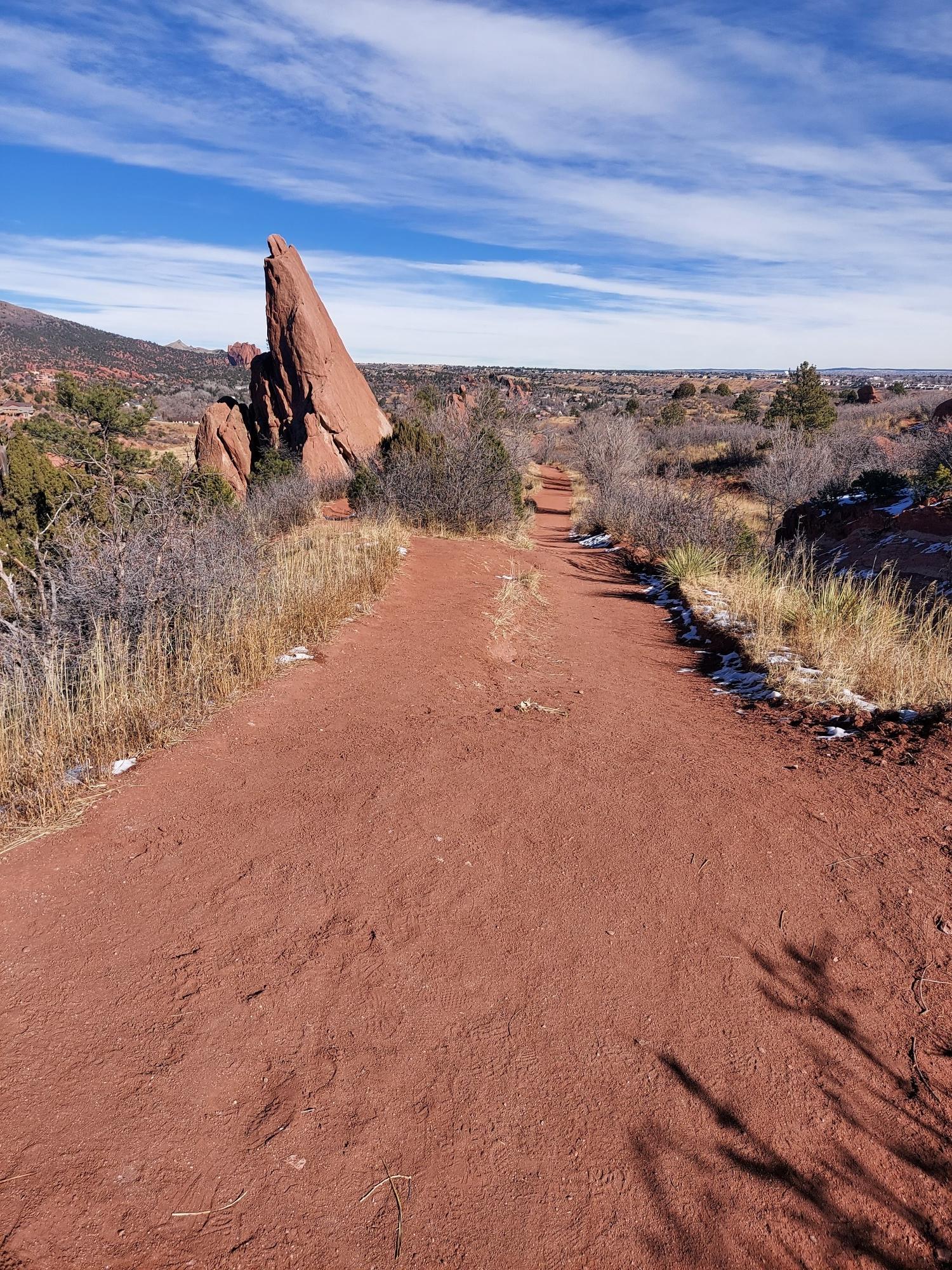 A red dirt path leads to a tall, pointed red rock under a blue sky.