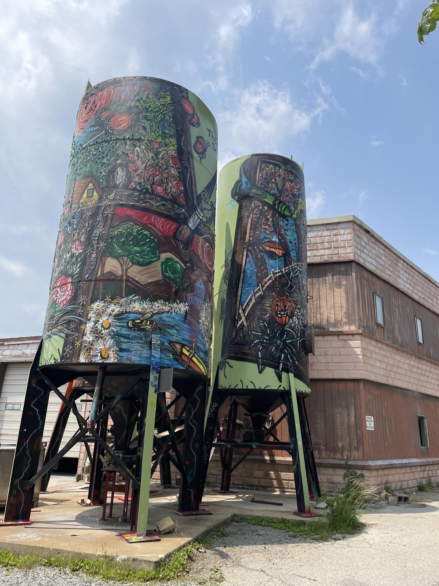 Two large, colorful, mural-painted silos stand under a partly cloudy sky.