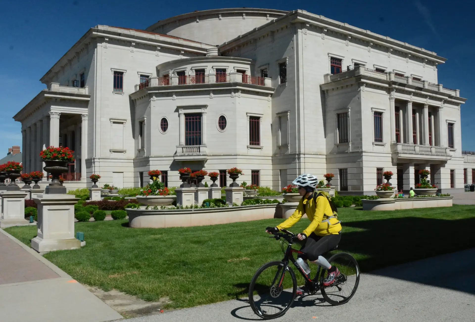 Cyclist in yellow jacket rides past a grand, light-colored building on a sunny day.