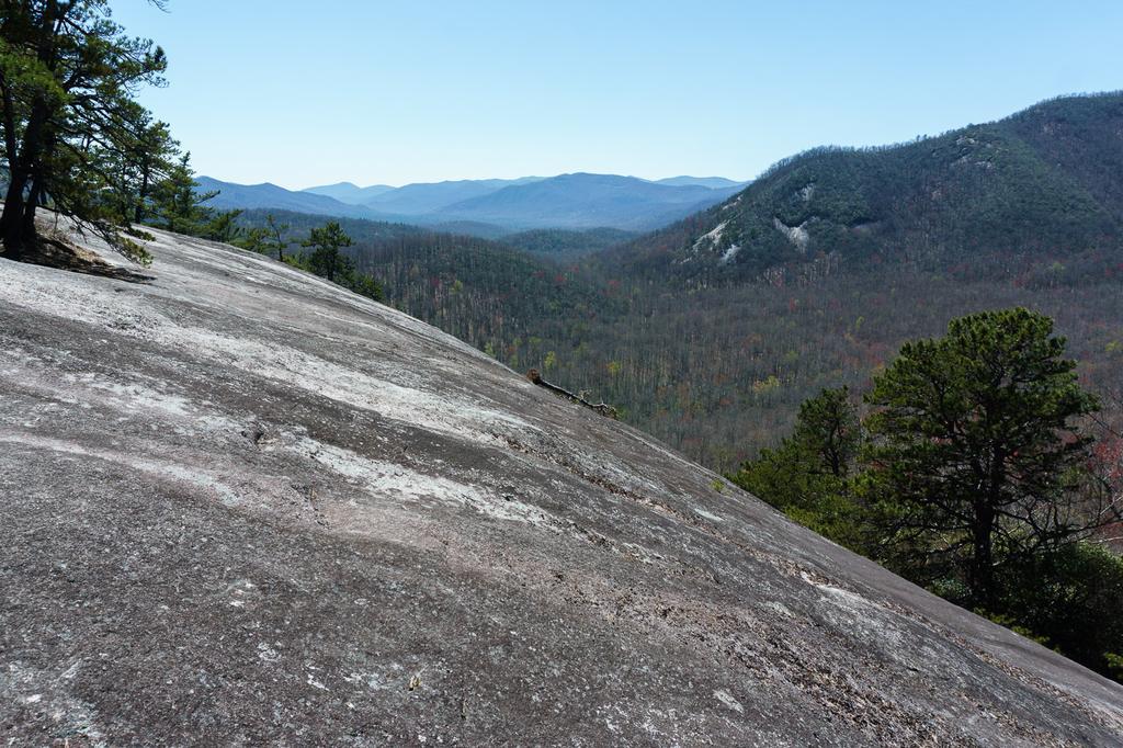 A large, sloping rock face overlooks a vast, sunny, forested mountain valley.