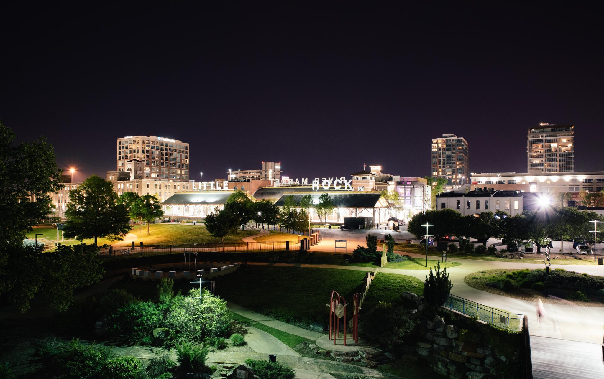 Illuminated city buildings and a green park at night with blurry figures.