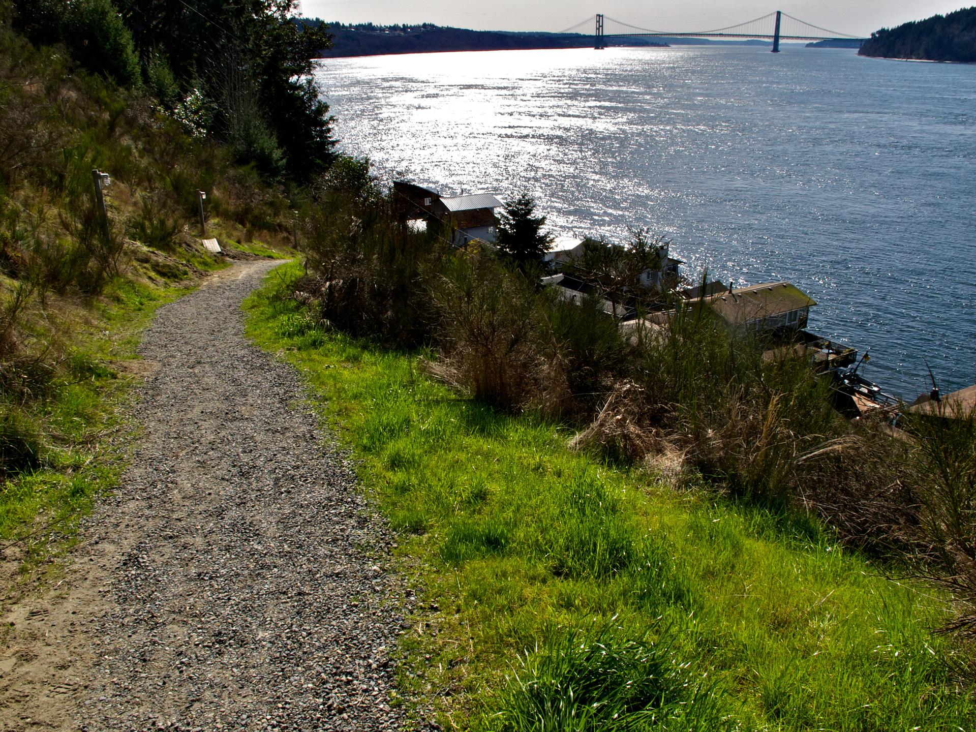 Gravel path on a sunny hillside overlooking sparkling water and a distant bridge.