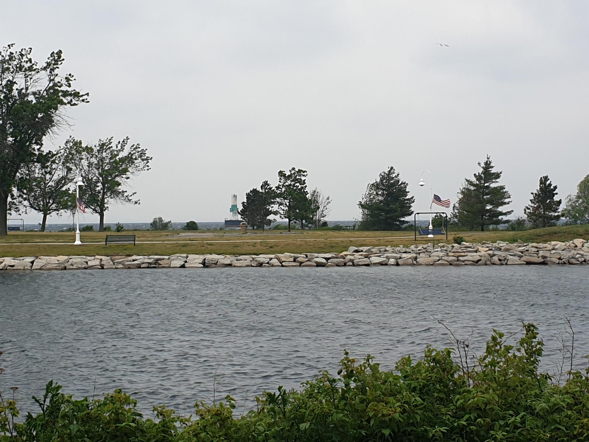 Calm water, rocky shore, and a park with trees under an overcast sky.