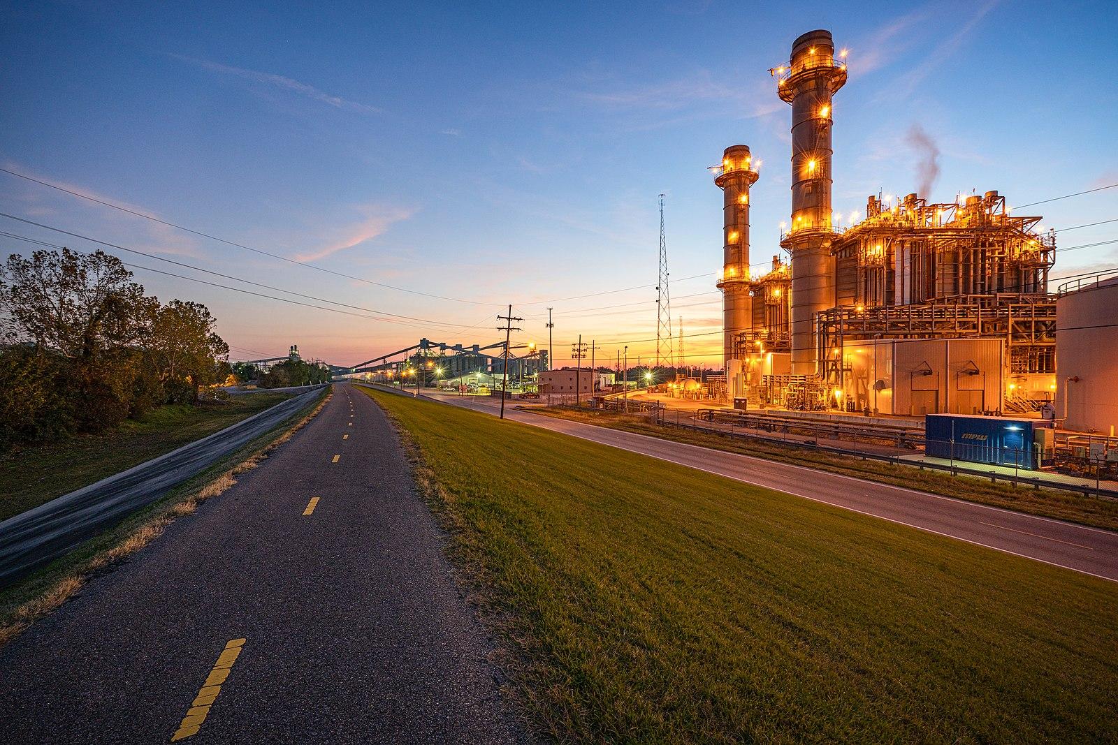 Industrial power plant illuminated at dusk beside a road and grassy area.