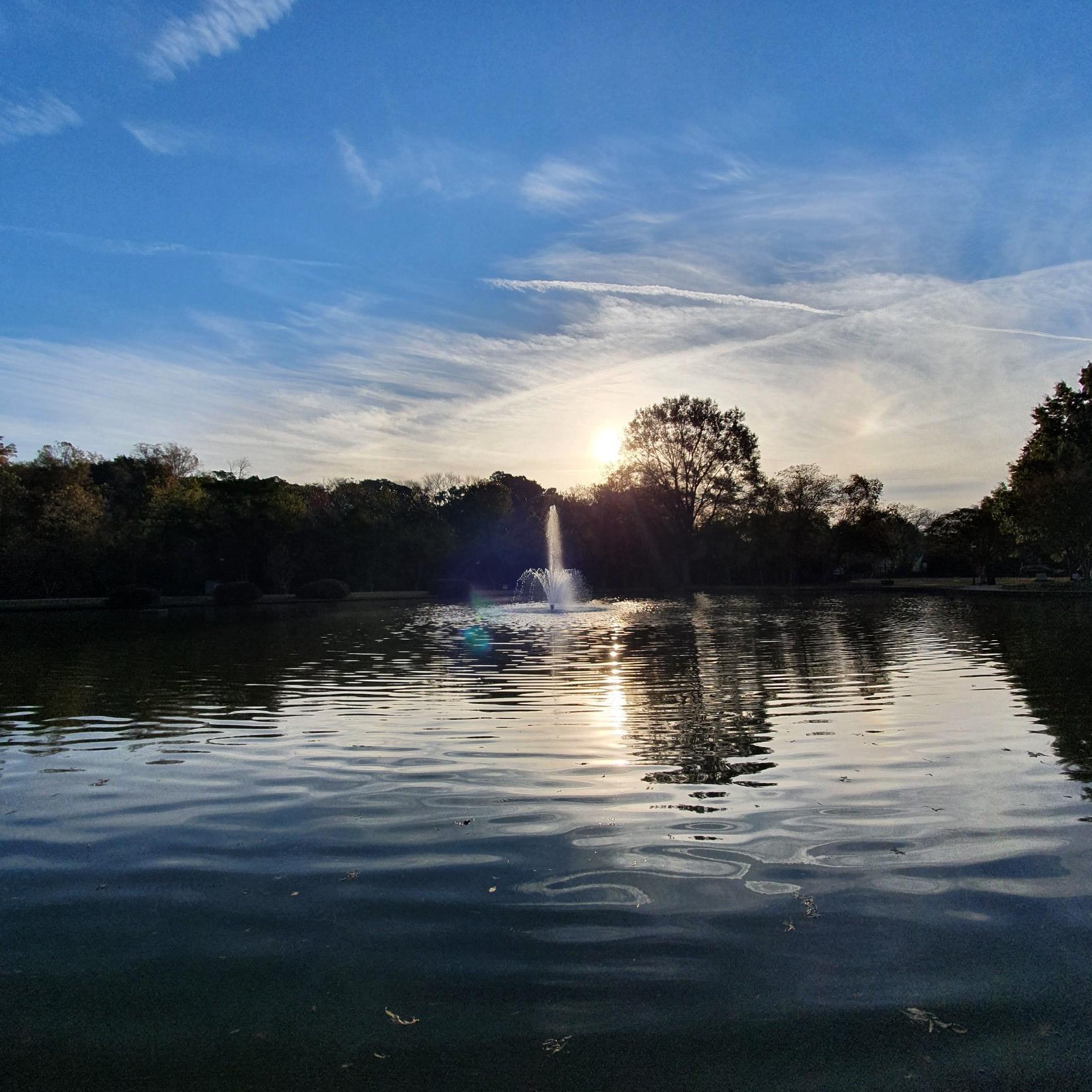 Sunset over a lake with a fountain, reflecting golden light on water.
