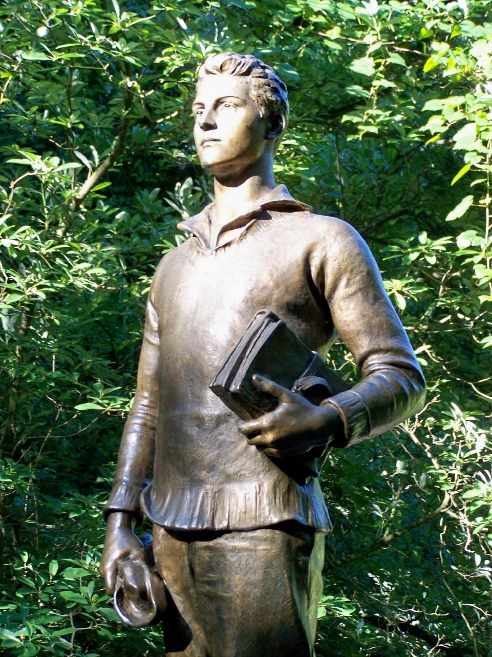 Sunny bronze statue of a young man with a book amidst green trees.