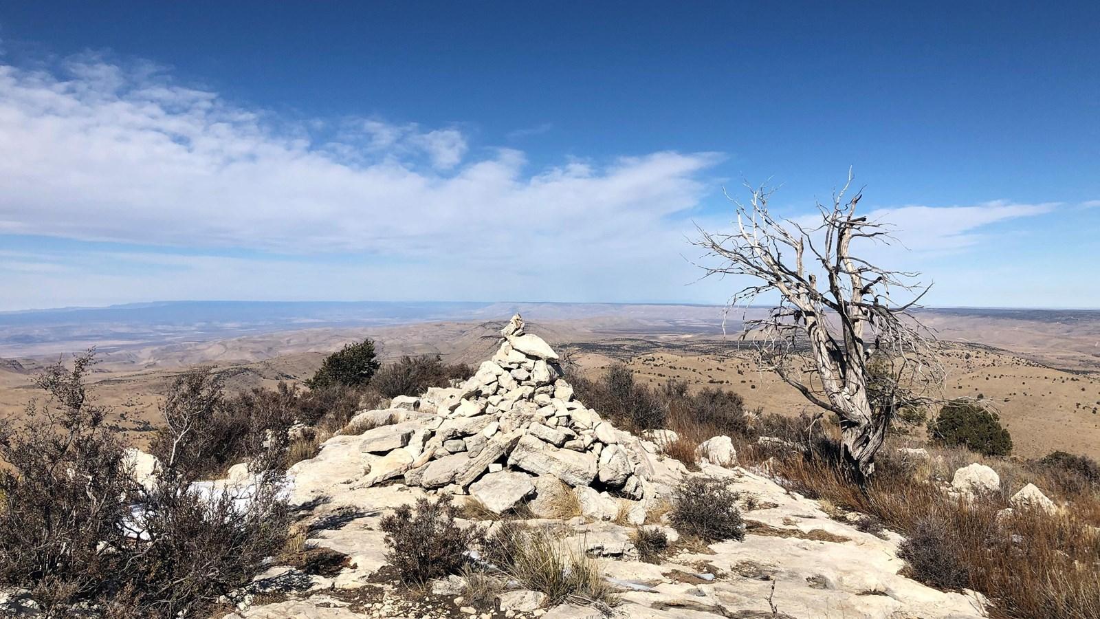 Stone cairn and dead tree on a rocky peak overlooking an arid landscape.