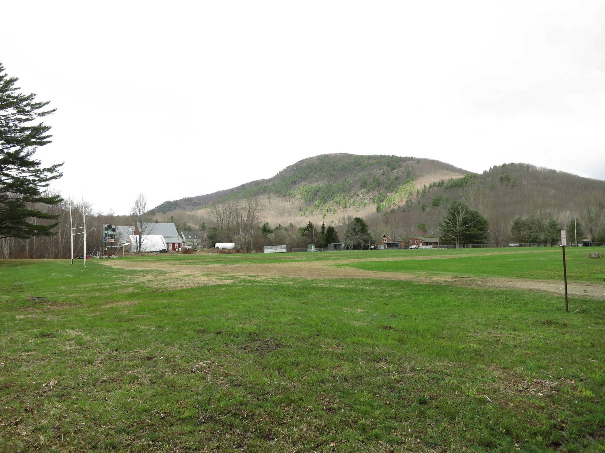 Grassy field with distant buildings and a forested mountain under an overcast sky. - Round the Mountain Trail